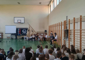 Uczniowie siedzący na sali gimnastycznej uczestniczą w apelu z okazji Święta Niepodległości, z pocztem sztandarowym i dekoracją biało-czerwoną na scenie.