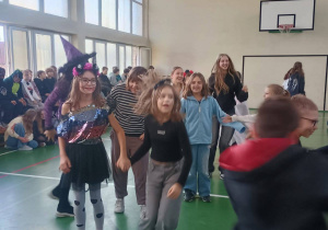 Grupa uczniów tańczy na sali gimnastycznej podczas dyskoteki halloweenowej; niektóre dzieci mają przebrania, a w tle siedzą inni uczniowie.