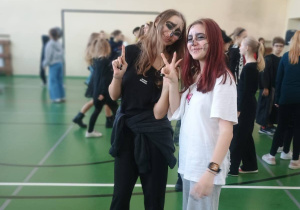 Dwie uczennice klasy 8 z halloweenowym makijażem pozują do zdjęcia na sali gimnastycznej, w tle inni uczniowie podczas szkolnej zabawy.