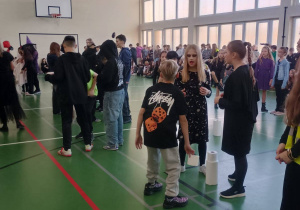 Uczniowie przebrani na Halloween biorą udział w zabawie na sali gimnastycznej, obok nich stoją rolki papieru toaletowego używane w konkursie.