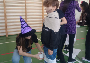 Dziecko owija kolegę papierem toaletowym podczas halloweenowego konkursu na sali gimnastycznej; oboje mają stroje i makijaże halloweenowe.