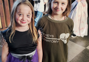 Dwie młodsze uczennice w halloweenowych strojach pozują na szkolnym korytarzu; jedna w czarno-fioletowym kostiumie z tiulem, druga w oliwkowej bluzie, w tle inne dzieci w przebraniach.