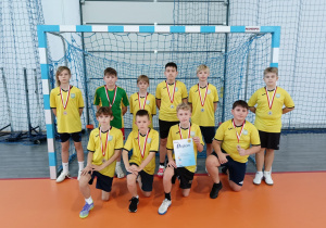 Grupa dziesięciu uczniów SP 13 w Pabianicach w żółtych strojach sportowych pozuje z medalami i dyplomem przed bramką piłki ręcznej na hali sportowej.
