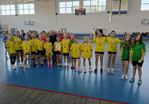 Dziewczęta z SP 13 w Pabianicach w żółtych koszulkach stoją na hali sportowej z medalami i dyplomem po zakończonych zawodach w piłce ręcznej.