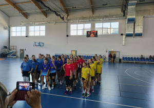 Drużyny dziewcząt w różnych kolorach strojów ustawione w kolumnach na hali sportowej; z prawej strony idą zawodniczki SP 13 w Pabianicach w żółtych koszulkach.