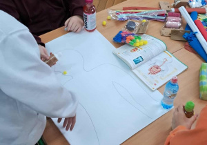 Uczniowie przy ławce rysują sylwetkę człowieka na arkuszu papieru, przygotowując się do wykonania modelu układu pokarmowego z różnorodnych materiałów.