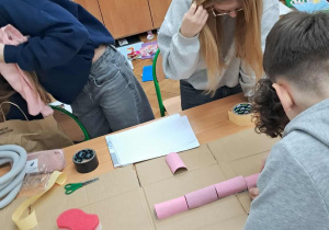 Grupa uczniów pochylona nad ławką układa elementy modelu układu pokarmowego na dużym kartonie w pracowni biologicznej.