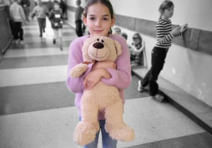 „Dziewczynka na szkolnym korytarzu trzyma w ramionach swojego pluszowego misia, wyróżniona kolorem na czarno-białym tle.”