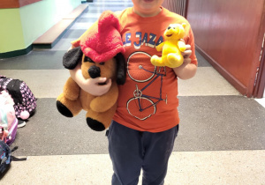 „Uczeń w pomarańczowej koszulce prezentuje dwa pluszaki – psa i żółtego misia – podczas szkolnego Dnia Pluszowego Misia.”