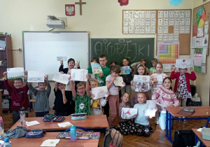 Klasa 2b stoi w sali przed tablicą z napisem „Andrzejki”, a dzieci trzymają nad głowami prace plastyczne inspirowane andrzejkowymi wróżbami z wosku.