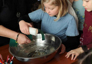 Dziewczynka z klasy 2a pochyla się nad metalową miską i przelewa wosk z świecy przez kartonowy otwór, otoczona rówieśnikami w czasie andrzejkowej zabawy.