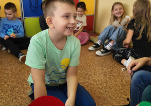 Uczeń klasy 1b siedzi na podłodze z czerwonym kapeluszem do losowania karteczek w czasie andrzejkowych zabaw.