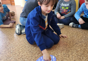 Uczniowie klasy 1b siedzą w kręgu na podłodze, chłopiec na pierwszym planie wskazuje palcem kartonowe serce z przyklejonymi karteczkami – bierze udział w andrzejkowej wróżbie z imionami.