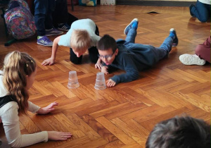 Uczniowie klasy 2a biorą udział w wyścigu na brzuchach po parkiecie, ścigając się do plastikowych kubeczków podczas andrzejek.