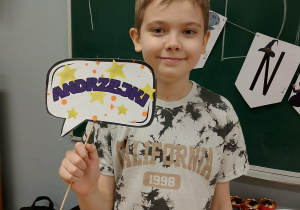 Uczeń 6a pozuje z dekoracyjną chmurką „ANDRZEJKI” na tle tablicy z napisem i stołu ze słodyczami.