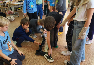 Uczniowie klasy 1b stoją i kucają wokół rzędu ustawionych butów, biorąc udział w wróżbie andrzejkowej.