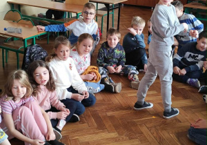 Grupa dzieci z klasy 2a siedzi na parkiecie w kole, przed nimi znajduje się metalowa miska z wodą do andrzejkowych zabaw.