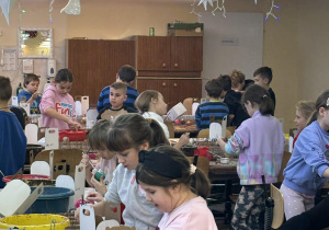 Uczniowie klas drugich dekorują bombki przy długich stołach w świątecznie udekorowanej hali fabryki bombek.