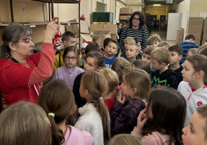 Dzieci z klas 2a i 2b słuchają pracownicy fabryki, która prezentuje im gotową świąteczną bombkę w czasie zwiedzania zakładu.