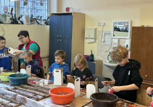 Grupa uczniów z klas 2a i 2b dekoruje bombki, a nauczycielka pomaga im przy stole z farbami i brokatem.