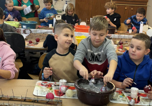 Chłopcy z klas drugich zanurzają bombki w misce z brokatem podczas warsztatów w fabryce bombek.