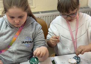 Dwie dziewczynki z młodszych klas ozdabiają zielone bombki białą farbą podczas warsztatów świątecznych w fabryce bombek.