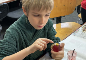 Chłopiec z klasy drugiej posypuje czerwoną bombkę złotym brokatem nad miską w czasie zajęć w fabryce bombek.