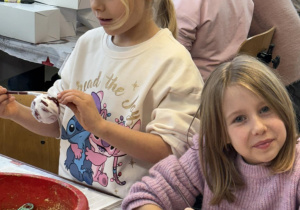 Dwie dziewczynki malują bombki i korzystają z pojemników z brokatem podczas warsztatów plastycznych w fabryce bombek.
