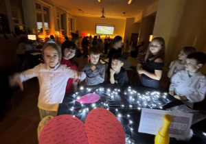 Dzieci podczas andrzejkowej zabawy w świetlicy stoją przy stoliku z lampkami, a na pierwszym planie widoczne jest różowe serce z imionami używane do wróżb miłosnych.