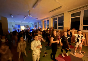 Grupa dzieci tańczy na andrzejkowej dyskotece w świetlicy, w przyciemnionej sali oświetlonej dekoracyjnymi lampkami rozmieszczonymi wzdłuż okien.