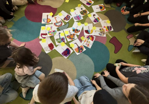 Grupa uczniów w świetlicy siedzi w kręgu na dywanie i ocenia kolorowe parasole z bibuły wykonane w ramach listopadowych zajęć plastycznych.