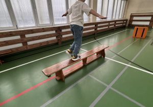 Chłopiec z rozłożonymi rękami idzie po ławce gimnastycznej na sali, ćwicząc równowagę w czasie olimpiady świetlicowej.
