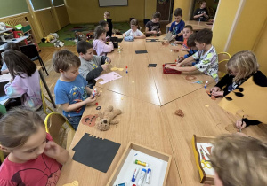 Uczniowie pracują przy stołach nad papierowymi misiami, a w tle na ekranie widać film o pluszowych misiach – zajęcia z okazji Dnia Pluszowego Misia.