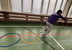 Uczeń skacze przez kolorowe koła ułożone na podłodze sali gimnastycznej w czasie świetlicowego toru przeszkód.