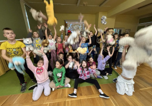 Grupa dzieci w świetlicy skacze i jednocześnie wyrzuca w górę pluszowe misie, radośnie obchodząc Dzień Pluszowego Misia.