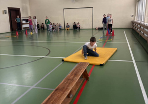Dziewczynka pokonuje tor przeszkód na żółtej macie podczas Świetlicowej Olimpiady Sportowej, a grupa dzieci siedzi z boku sali gimnastycznej i jej kibicuje.