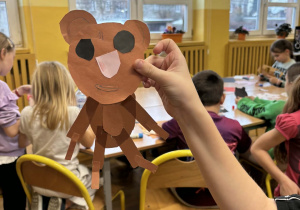 Zbliżenie na papierowego misia trzymanego przez ucznia, w tle dzieci siedzą przy stolikach i tworzą kolejne misiowe prace plastyczne w świetlicy.