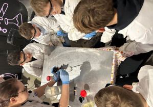 Prowadząca wykonuje doświadczenie z odczynnikiem, a uczniowie w goglach i fartuchach skupieni patrzą na szalkę podczas zajęć w Experymentarium.