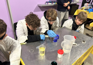 Uczniowie klasy 7a w fartuchach mieszają niebieskie roztwory w zlewkach, samodzielnie wykonując doświadczenia chemiczne na warsztatach.