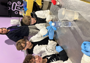 Chłopcy z klasy 7a przeprowadzają doświadczenie z balonem i reagującymi substancjami, a edukator z Experymentarium obserwuje ich pracę