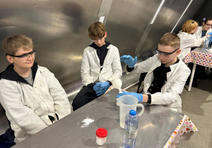 Uczeń klasy 7a przygotowuje doświadczenie chemiczne z użyciem balonu i roztworu, pozostali uczestnicy obserwują przebieg eksperymentu.