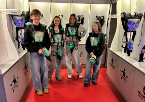 Uczniowie klasy 7a w kamizelkach z sensorami i z pistoletami laserowymi pozują przed wejściem na arenę Laser Games podczas klasowych mikołajek.