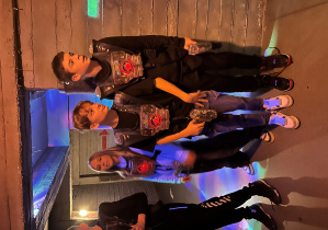 Troje uczniów 7a w czerwono podświetlonych kamizelkach laserowych czeka przy wejściu na arenę Laser Games przed kolejną rundą.