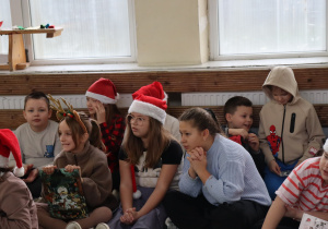 Uczniowie klas I–VI w mikołajkowych strojach siedzą na sali gimnastycznej podczas Mikołajek.