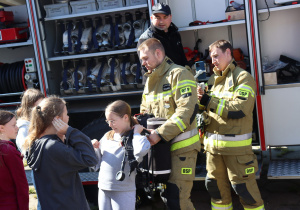 Na zewnątrz, przy otwartym wozie strażackim, strażacy z OSP pokazują dzieciom sprzęt gaśniczy. Dziewczynka w szarym stroju przymierza aparat powietrzny z pomocą strażaka. W tle grupa dzieci przygląda się prezentacji, a za nimi widoczne są węże strażackie i inne wyposażenie pojazdu.