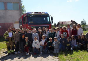 Grupowe zdjęcie dużej grupy dzieci i młodzieży wraz ze strażakami na tle czerwonego wozu strażackiego marki Volvo. Jeden ze strażaków trzyma w rękach dyplom. Zdjęcie wykonane na zewnątrz, przy budynku, w słoneczny dzień. W tle widoczne domy i drzewa.