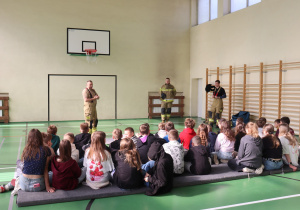 Zdjęcie przedstawia grupę uczniów siedzących na materacach w sali gimnastycznej, zwróconych w stronę trzech strażaków w mundurach OSP. Strażacy stoją pod ścianą z tablicą do koszykówki, prowadząc pogadankę lub prezentację. W tle widać drabinki gimnastyczne i okna wpuszczające światło dzienne.