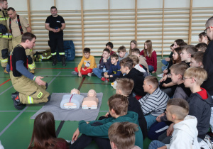 Strażak w ubraniu ochronnym OSP klęczy na sali gimnastycznej przed dwiema fantomami do resuscytacji krążeniowo-oddechowej (RKO), tłumacząc zasady udzielania pierwszej pomocy. Przed nim siedzi duża grupa dzieci w wieku szkolnym, uważnie słuchająca instrukcji. W tle widać innych strażaków oraz drabinki gimnastyczne na ścianie.