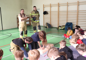Na tym zdjęciu widać, jak uczennica ćwiczy resuscytację krążeniowo-oddechową (RKO) na fantomie, pod czujnym okiem strażaka z OSP. Pozostali uczestnicy zajęć z uwagą obserwują przebieg ćwiczenia. W tle stoją strażacy, którzy wspierają prowadzenie szkolenia. Tego rodzaju zajęcia są nie tylko edukacyjne, ale również motywujące dla dzieci – pozwalają poczuć się pewniej w sytuacjach zagrożenia życia i uczą praktycznych umiejętności, które mogą uratować komuś życie.