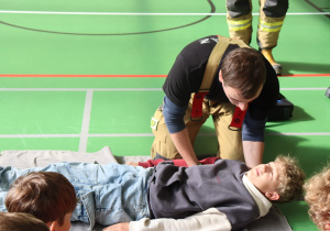 Na zdjęciu widać strażaka ochotnika, który prowadzi praktyczne szkolenie z zakresu udzielania pierwszej pomocy. Jeden z uczniów leży na macie, a strażak pokazuje na nim odpowiednie techniki — najprawdopodobniej ocenę przytomności lub oddechu. Pozostali uczniowie uważnie obserwują, co wskazuje na duże zainteresowanie i zaangażowanie w zajęcia.